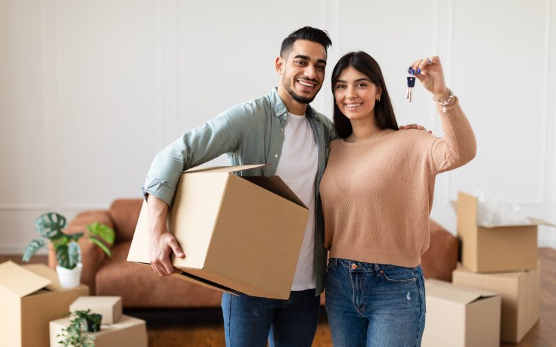 House Ownership. Young Couple Showing Keys And Holding Cardboard Box, Cheerful Guy And Lady Hugging After Moving In New Apartment Standing In Living Room. Insurance, Real Estate, Mortgage Concept